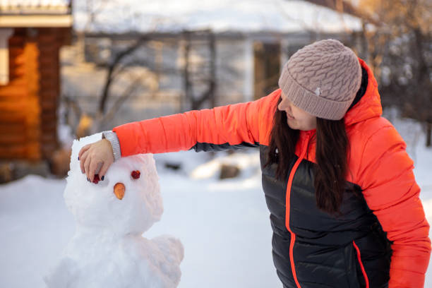 istock Middle aged female making face of snowman with improvised means in daytime while having walk on backyard with trees, houses and bright sun light in background. Parents spending time with children 1365376399