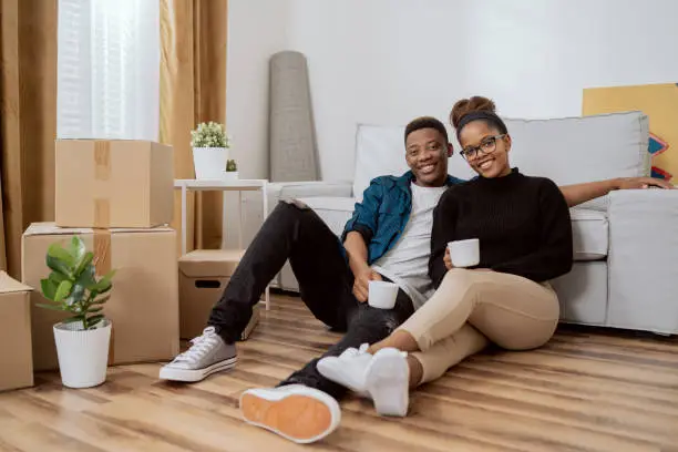 Photo of Smiling couple sitting on the floor relaxing after moving in, drinking coffee in their new apartment, around them boxes with unpacked things, the husband embraces his wife