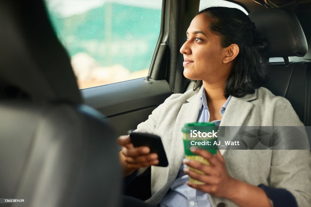 Woman travelling by the car Woman sitting in backseat of her car holding coffee and mobile looking outside car window Crowdsourced Taxi Stock Photo