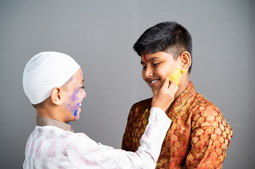 Multiethnic religious kids celebrating holi by applying colors to face on gray background - concept of cultural festivals, friendship and unity in diversity.