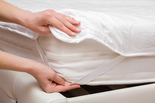 Young adult woman hands putting white cover with elastic band on mattress corner. Closeup. Front view. Regular bed linen change.