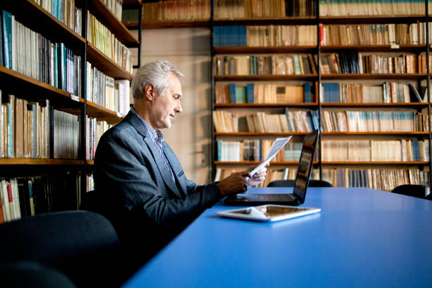 University senior professor in library looks at student statistics stock photo Teacher, Professor, Senior Adult, University,Library,Book,Senior,Men,60-69 Years,40-49 Years,50-59 Years,Photography professor business classroom computer stock pictures, royalty-free photos & images