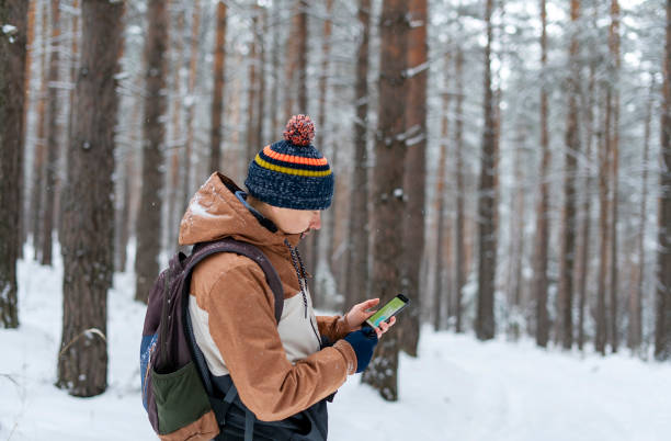 istock Young man in knitted hat with backpack looking at map on smartphone while hiking in winter forest using modern technology navigation 1365393018