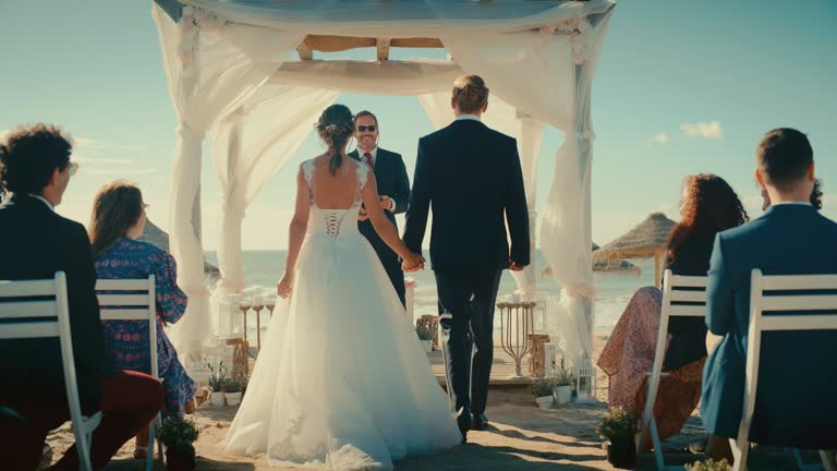 Beautiful Bride in White Wedding Dress and Handsome Groom in a Traditional Black Suit Going Down the Aisle at an Outdoors Ceremony Venue Near the Sea with Happy Multiethnic and Diverse Friends.