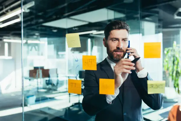 Photo of businessman in reflection at a glass task board, a man talking on the phone, hanging plans on colored paper stickers, plans for the near future