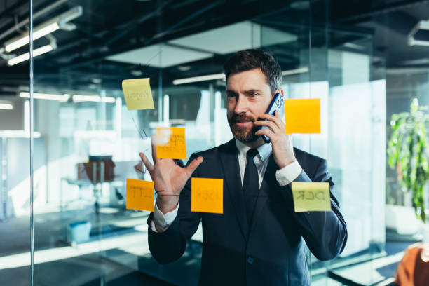 istock businessman in reflection at a glass task board, a man talking on the phone, hanging plans on colored paper stickers, plans for the near future 1365400249