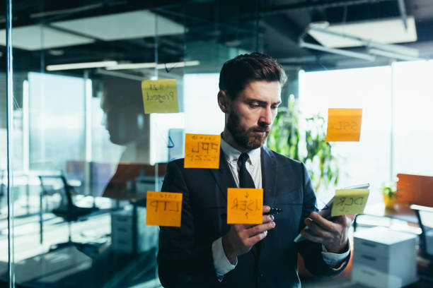 istock Successful businessman rewrites a task on a glass board with colored stickers, from phone and tablet 1365400324