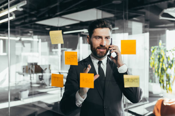 istock businessman in reflection at a glass task board, a man talking on the phone, hanging plans on colored paper stickers, plans for the near future 1365400404