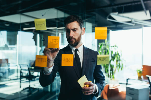 istock businessman in reflection at a glass task board, hangs plans on colored paper stickers, plans for the near future 1365401204