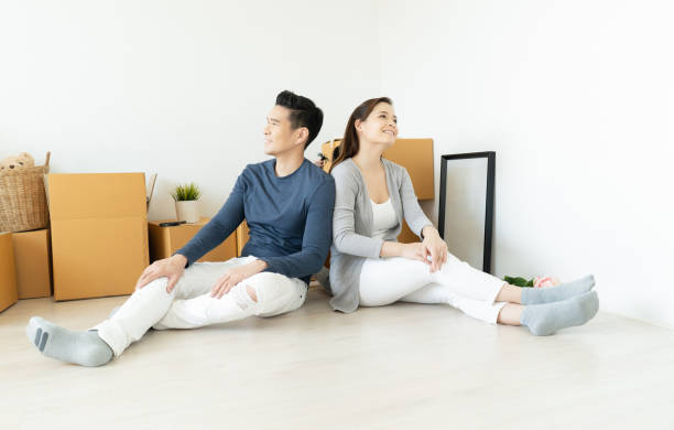 feliz joven pareja asiática durante la mudanza de casa sentada en el suelo en una nueva casa con caja de desempacar y mirando hacia otro lado. hogar, personas, mudanzas y concepto inmobiliario. - moving house physical activity real estate couple fotografías e imágenes de stock