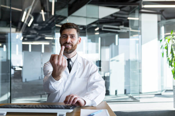 Handsome doctor man in coat and stethoscope showing middle finger, fuck you bad expression, provocation and rudeness. He shouts excited Handsome doctor man in coat and stethoscope showing middle finger, fuck you bad expression, provocation and rudeness. He shouts excited fighting stance stock pictures, royalty-free photos & images