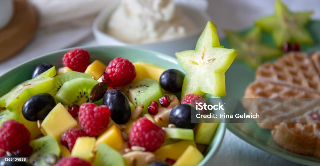 Fresh healthy fruit salad blue green cup on white background with carambola Green-blue bowl with fresh, healthy fruit salad on a white background with carambola, in the background a plate with waffles and a stack of fresh whipped cream Banana Stock Photo