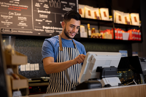 Portrait of a happy Latin American man working as a cashier at a cafe and placing an order in the computer