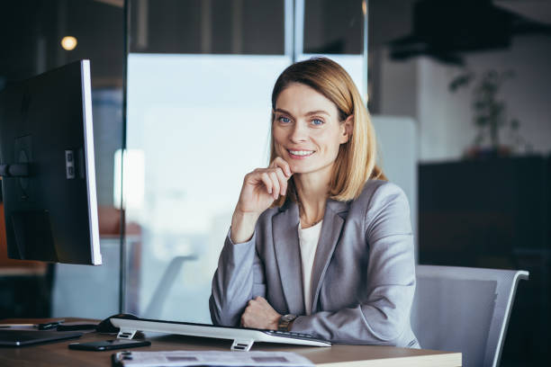 patronne d’affaires heureuse et prospère travaillant à l’ordinateur dans un bureau moderne regardant la caméra et souriant - business women manager looking at camera photos et images de collection