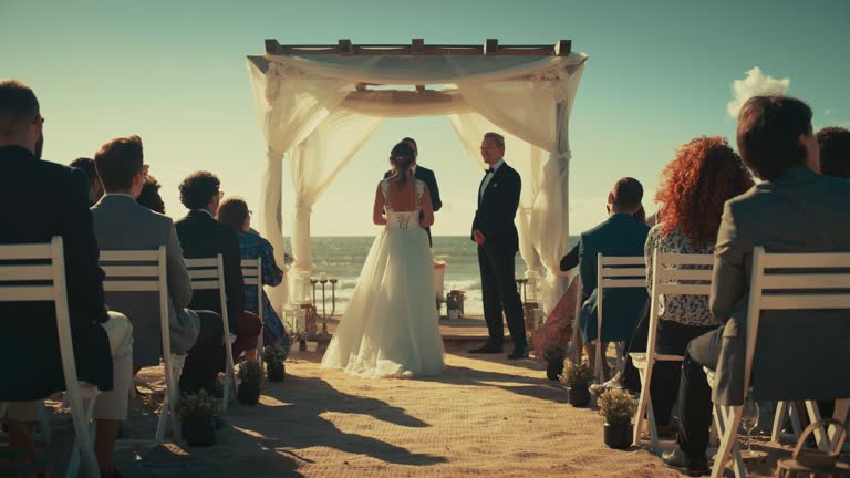 Beautiful Bride in Gorgeous White Wedding Dress Going Down the Aisle, while Groom Waits at an Outdoors Ceremony Venue Near the Sea with Happy Multiethnic and Diverse Friends.