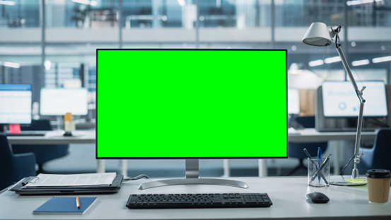 Monitor de computadora de escritorio con maqueta de pantalla verde Chroma Key Display de pie en el escritorio en la oficina de negocios moderna. En la pared de vidrio de fondo con la oficina de la gran ciudad. photo