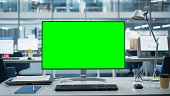 istock Desktop Computer Monitor with Mock Up Green Screen Chroma Key Display Standing on the Desk in the Modern Business Office. In the Background Glass Wall with Big City Office. 1365412419