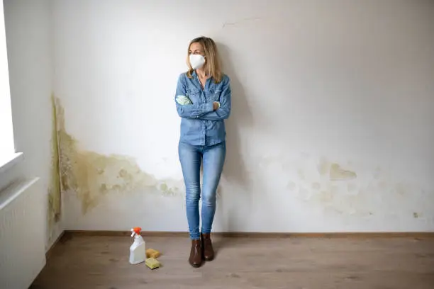 Photo of blonde young woman standing in front of white apartment wall with mold on it frustrated and desperate