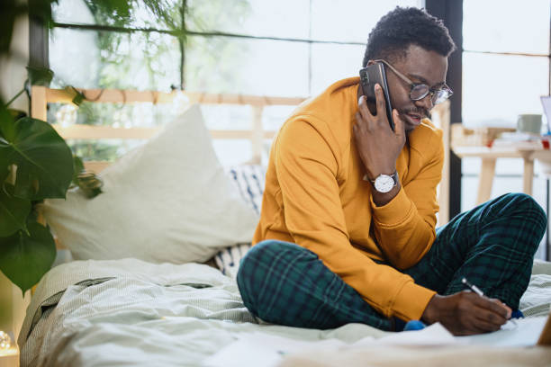 istock Male student talking on the phone while sitting on the bed in his bedroom studying 1365414959