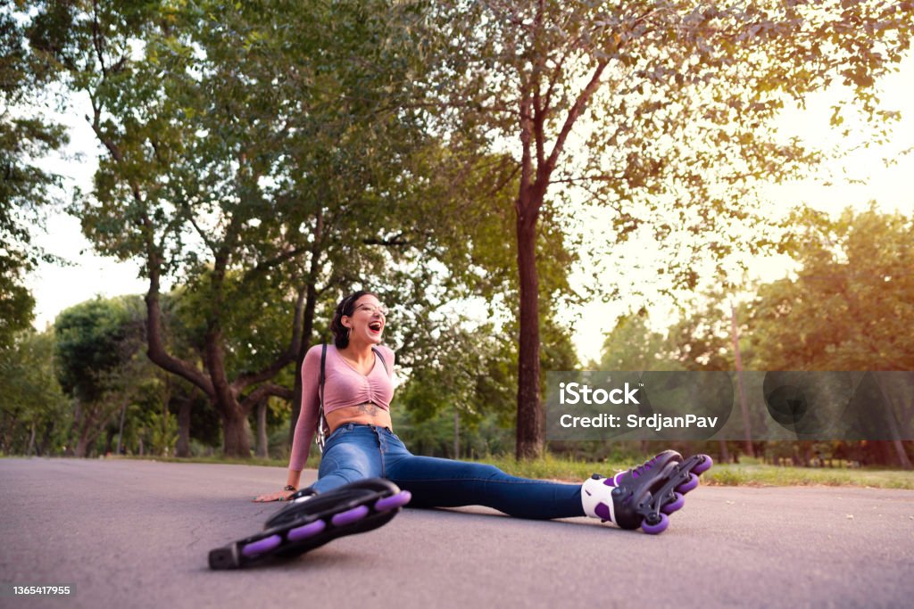 Young female skater laughing while sitting on the road after the fall Young Caucasian woman with her roller skates on, laughing while sitting on the road after the fall. Falling Stock Photo