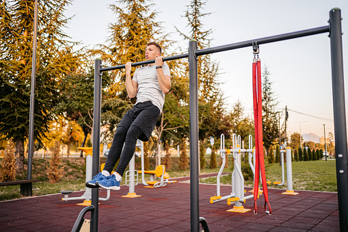 A handsome young man doing chin-ups on a bar outdoors in a public park.