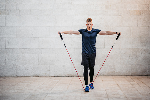 A handsome young man training outdoors by doing lateral raises with a resistance band.