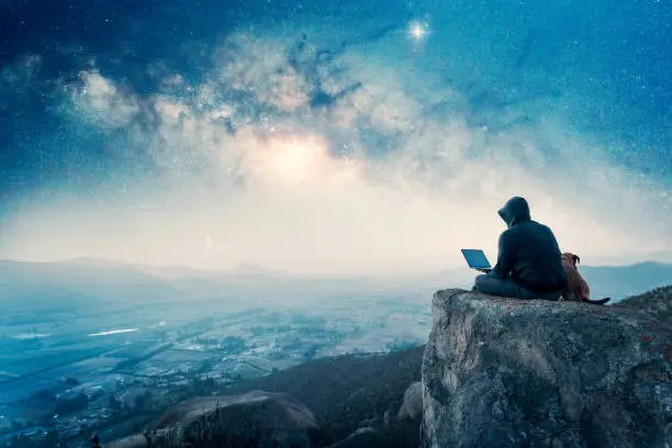 Photo of silhouette of a person sitting on the top of the mountain with a laptop and a dog, over the city and Milky Way background