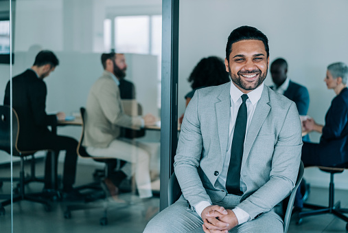 Shot of a handsome smiling businessman standing in front of his team in the office. Portrait of successful businessman sitting in office chair with his colleagues working in background. Multi-ethnic group of corporate businesspeople on a meeting in board room.