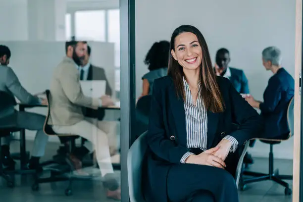 Shot of a beautiful smiling businesswoman standing in front of her team in the office. Portrait of successful businesswoman sitting in office chair with her colleagues working in background. Multi-ethnic group of corporate businesspeople on a meeting in board room.