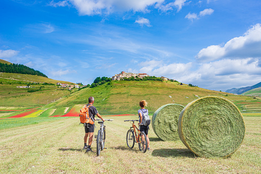 Couple cycling mtb in blooming cultivated fields of Castelluccio di Norcia highlands, famous colourful flowering plain in the Apennines, Italy. Agriculture of entil crops and red poppies.