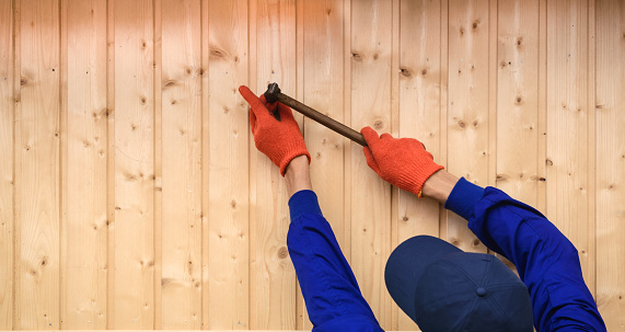 Young man contractor worker is holding a hammer and drive a nail in to a wood wall from boards in construction site. Copy space for you text