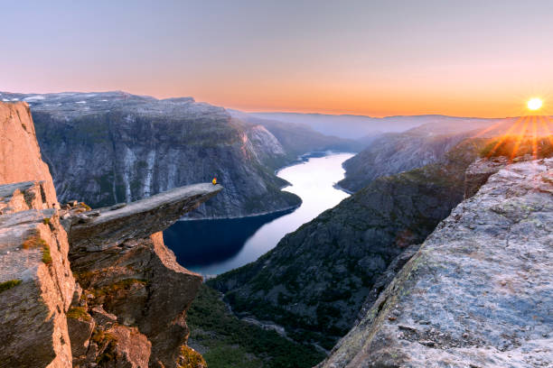 ein mann sitzt, während er seine arme in die luft wirft, auf der klippe des berges trolltunga, drängt sich über ringedalsvatnet und beobachtet den sonnenuntergang in den verschneiten norwegischen bergen in der nähe von odda, rogaland, norwegen - fjord stock-fotos und bilder