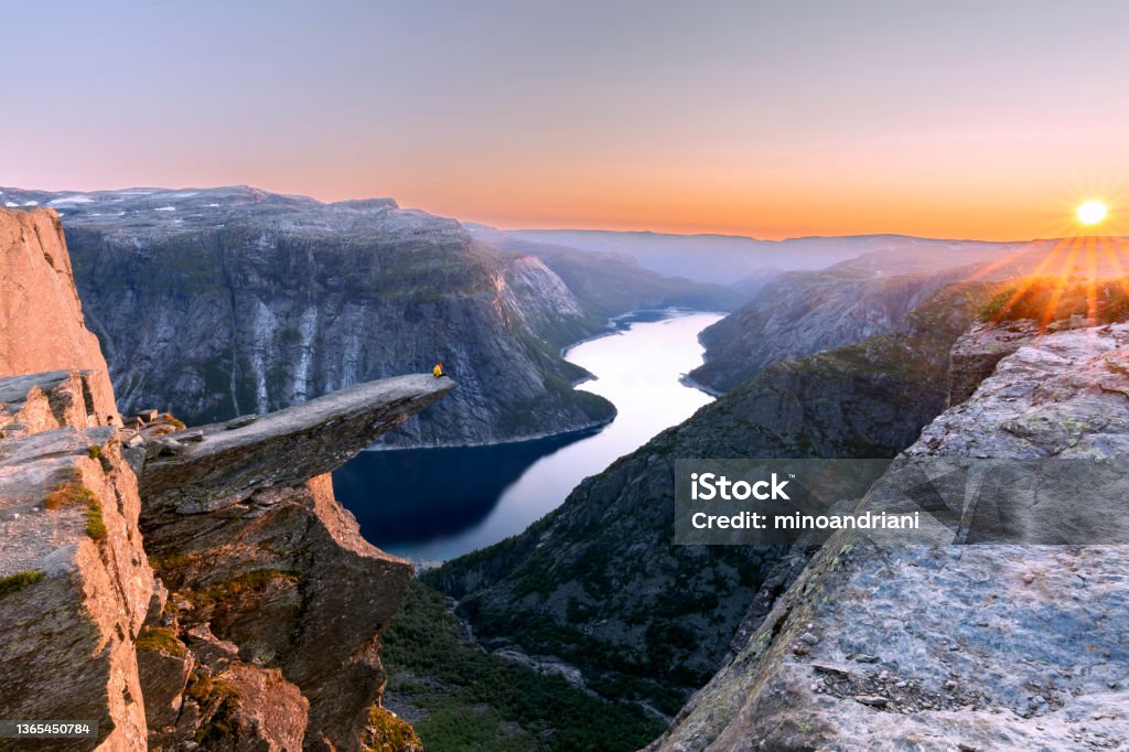 A man sits while throwing his arms in the air on the mountain's cliff edge of Trolltunga throning over Ringedalsvatnet watching the sunset in the snowy Norwegian mountains near Odda, Rogaland, Norway Norway Stock Photo