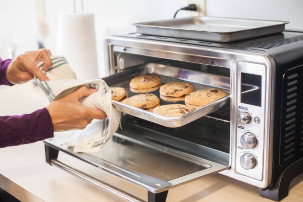 Baking Cookies in a toaster oven A female hand using a towel to remove a small baking sheet full of chocolate chip cookies from a silver toaster oven. The scene is a brightly lit modern kitchen counter. baking sheet stock pictures, royalty-free photos & images