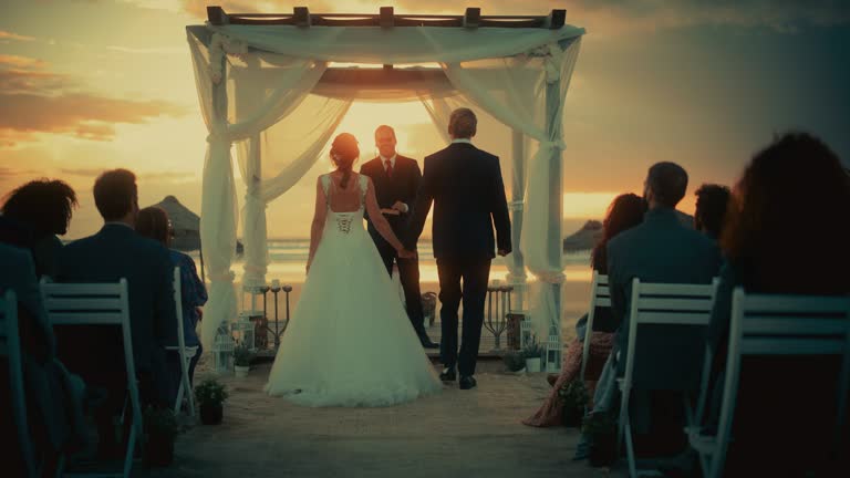Beautiful Bride in White Wedding Dress and Handsome Groom in Black Suit Going Down the Aisle at an Outdoors Ceremony Venue Near the Sea at Sunset. Happy Multiethnic Friends Celebrating Marriage.