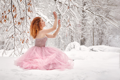 Winter fairy tale, a beautiful young woman in a luxurious lush pink dress enjoying in the snowy forest