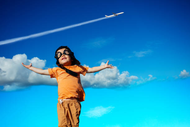 Little toddler boy in aviator hat and googles with happy lifted up hands over commercial jet plane in the sky