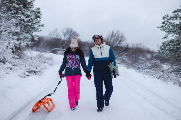 istock Beautiful young couple wearing colorful outfits holding hands while walking down a path with their snowboard and sled 1365496103