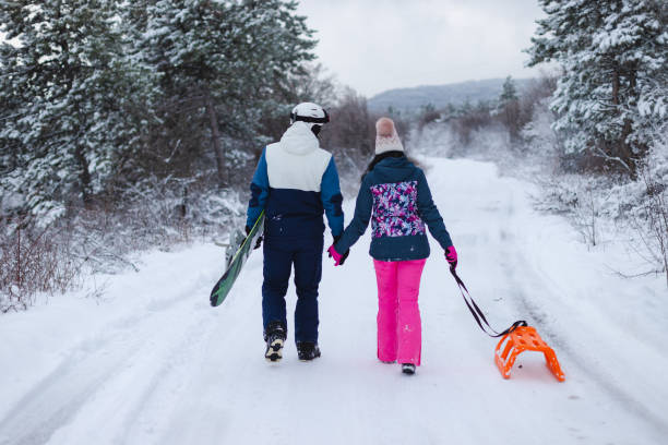 istock Beautiful young couple wearing colorful outfits holding hands while walking down a path with their snowboard and sled 1365496969