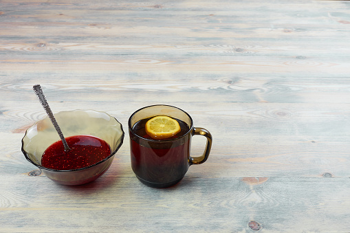 There is a mug of tea on a wooden table with a lemon wedge floating in it. Nearby is a vase of raspberry jam and a teaspoon in it.