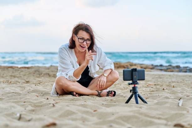 istock Middle aged woman sitting on beach with smartphone using video call 1365526687