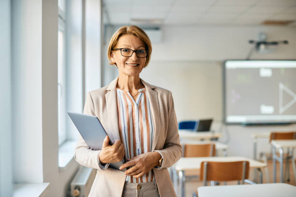 Portrait of smiling mature teacher with laptop in the classroom. Smiling mature teacher holding laptop while standing in the classroom and looking at camera. professor stock pictures, royalty-free photos & images