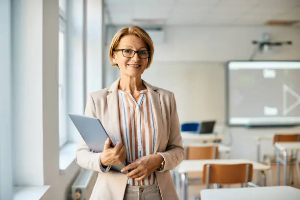 Smiling mature teacher holding laptop while standing in the classroom and looking at camera.