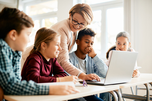 Happy elementary students and their teacher using laptop during computer class in the classroom.