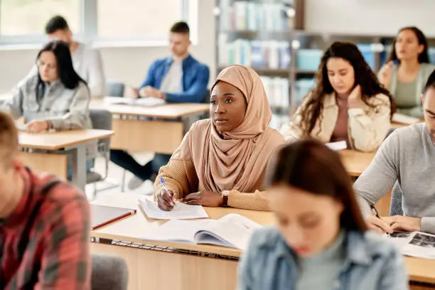 Female African American student in hijab writing during a class at university classroom.