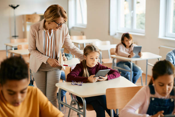 istock Schoolgirl using touchpad with teacher's assistance during a class in the classroom. 1365528029