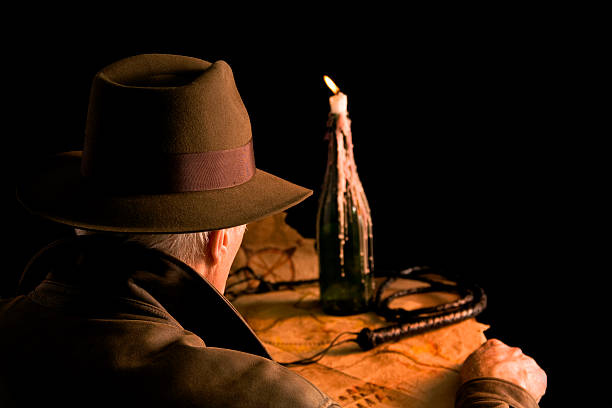 Indiana Jones style!  Man in brown leather coat and felt hat sitting at table with a map, his whip and a candle burning in a bottle.