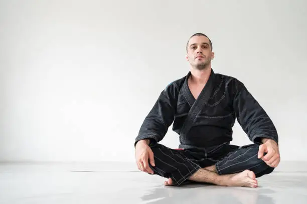 Front view of adult male athlete bjj brazilian jiu jistu black belt sitting in front of white wall in kimono gi with copy space looking to the camera