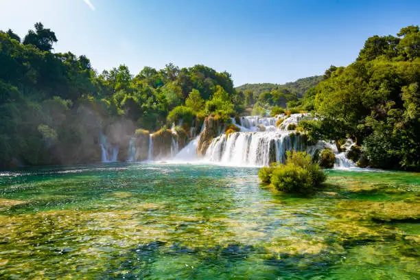 Photo of Waterfalls at Skradinski Buk, national nature park Krka, Croatia. Flowing water in beautiful nature, green plants and trees. Sunny summer day.