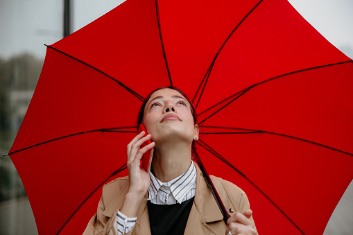 Attractive cheerful businesswoman using cellphone on the city street and holding a red umbrella while leaving the office.